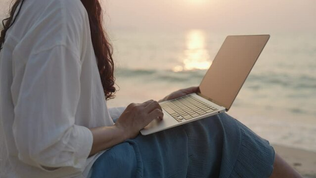 Young woman using laptop sitting work on the beach enjoying the sound of waves crashing on the sea shore at sunset