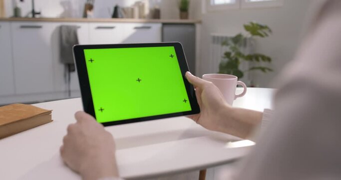 Women's hands holding large tablet with green screen on display. Woman sitting at table with cup of coffee and communicating via video call from tablet. Business call for work. Tablet with chromakey.