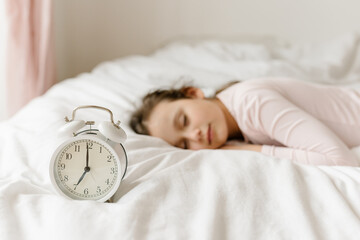 School child sleeps in bed. white alarm clock on foreground of bed and sleeping kid in interior of...