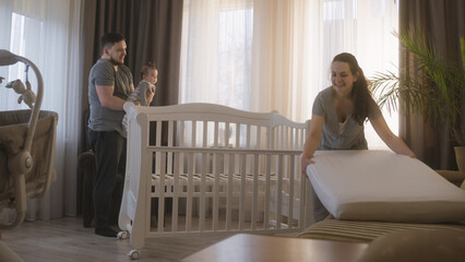 Loving mother puts mattress, blanket and child toys in baby crib. Caring father holds baby daughter...