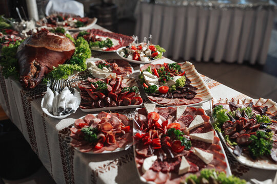 Buffet table at the wedding. An assortment of snacks on white plates standing on a Ukrainian-style tablecloth. Banquet service. Food, snacks with cheese, ham, prosciutto and greens.