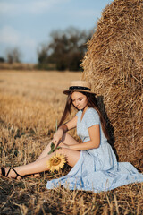 Naklejka na ściany i meble Portrait of a young girl. A girl in a blue dress and hat, holding a bouquet of sunflowers, against a background of hay bales. Long straight hair. Nice color. Summer