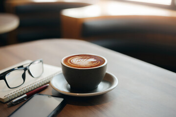 Close-up view, white cup of coffee with smartphone, notebook, pen and eye glasses on wooden table near bright window in cafe. Vintage light, blurred background