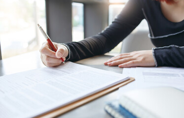 Cropped view of hands of young asian female in casual clothes working on desk using laptop while holding pen in her hands to review and sign new business deal, insurance contract or writing report