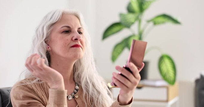 Mature woman fixes long grey hair examining face in small cosmetic mirror against houseplant. Lady checks aging skin condition at home slow motion