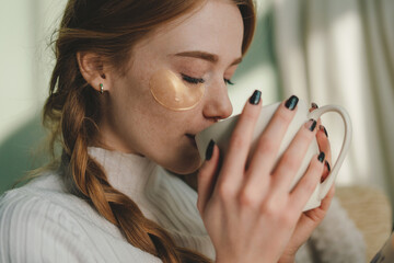 Close-up portrait ginger lady sitting on sofa and drinking coffee while wearing eye patches....