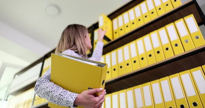 Secretary reaches over to upper shelf to take ring binder for paperwork. Organized yellow folders with archive materials in office slow motion