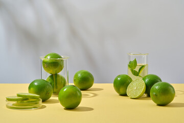 Front view of fresh limes and lab glassware decorated on white background with shadow leaves. Advertisement scene for cosmetics containing lime extract rich in vitamin c and minerals