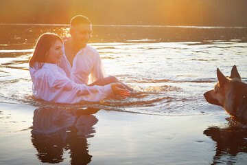 Beautiful adult couple has fun with bug dog shephers in nature in water of river or lake in summer evening at sunset. Guy and girl swim and relax with pet outdoors in clothes in white shirts and jeans