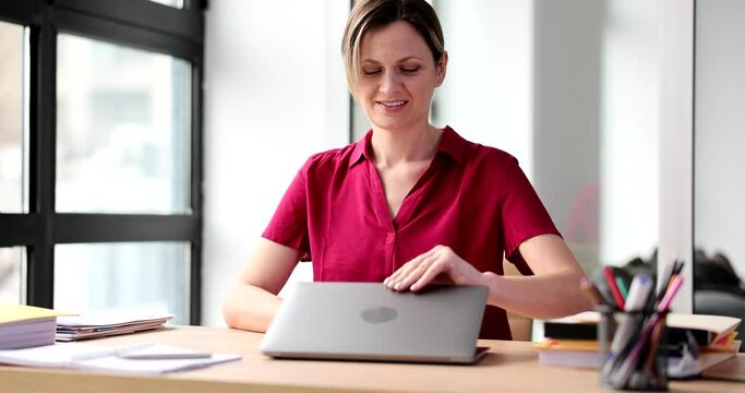 Positive woman opens laptop to start working on project. Blonde female employee in red blouse sits at table ready to use device slow motion