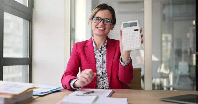 Happy woman shows big number on calculator after counting monthly profit of company. Emotional accountant in red jacket feels joy at work slow motion