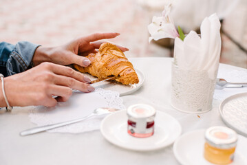 The girl holds a croissant with her hands that lies on a white plate on a table with a white tablecloth and cuts it with a table knife.