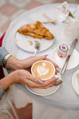 Women's hands hold a cup of coffee. A cut croissant lies on a table nearby against a blurred background. Table on the terrace of a summer cafe