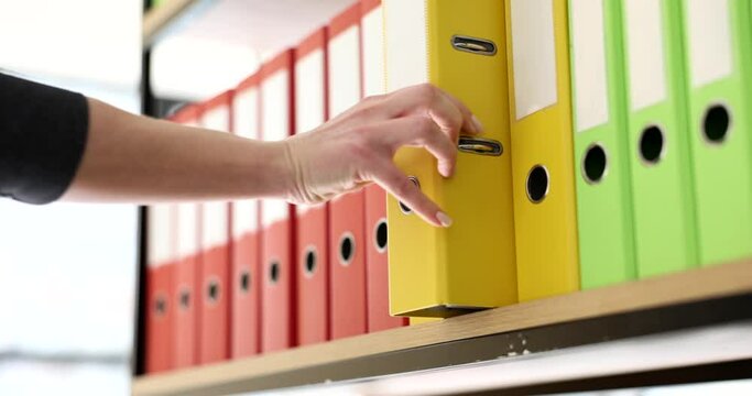 Secretary puts yellow folder with materials back on shelf of rack in office. Organized multi-colored ring binders with archive files slow motion