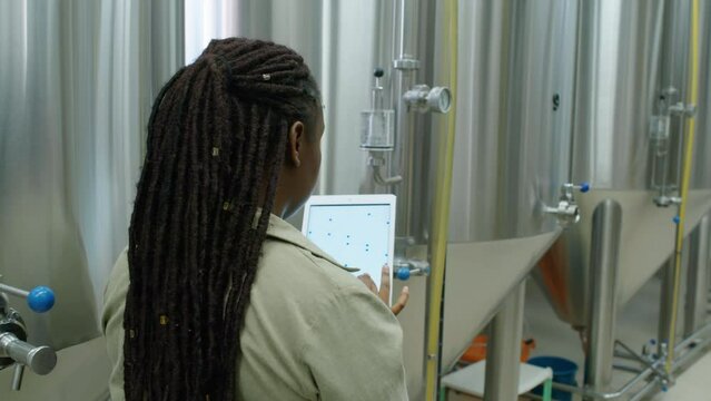 Medium rear over-shoulder shot of black female brewery supervisor with dreadlocks watching large brite beer tanks and completing checklist on tablet computer