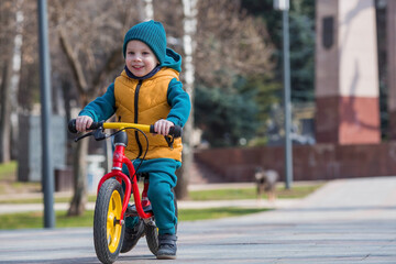 A cheerful little boy rides a bicycle outdoors. A happy child walks in the spring park. The baby is dressed in a fashionable yellow vest and turquoise jumpsuit.
