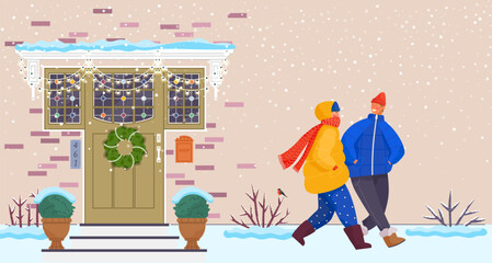 People wearing warm winter clothes walk down city street outside in cold season. Woman and man walking in snowy weather at house wall with entrance door decorated with Christmas wreath and decorations