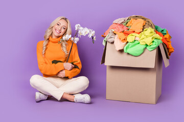 Full body portrait of cheerful pretty lady sit floor hold orchid flower pile stack clothes box isolated on purple color background