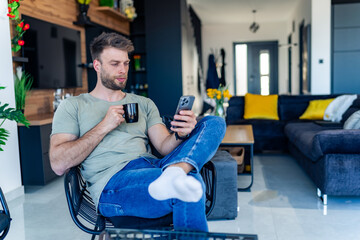 A good-looking man sits in a chair after a busy day, drinks coffee and uses the phone to read news on online portals.
