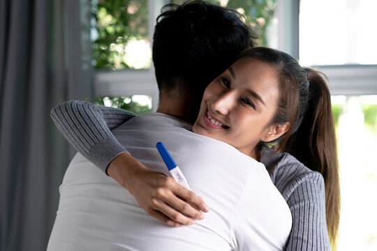 Happy young Asian woman hugging husband and holding positive pregnancy test in living room