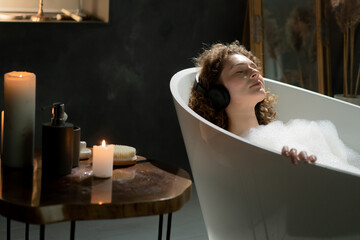 Photo of a beautiful woman who lies in the bath with her eyes closed and listens to music through wireless headphones. A young woman relaxes in a bath with aromatic candles and listens to music on