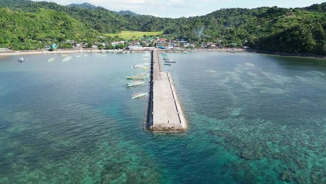 Cinematic Aerial Pullback Orbit View of Blue Outrigger Bangka Boat arriving at tropical village dock with serene seaside town and lush mountains in background. San Andres, Catanduanes.