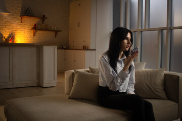 A young woman sits on a sofa in a dimly lit room and records a voice message on her phone. A...