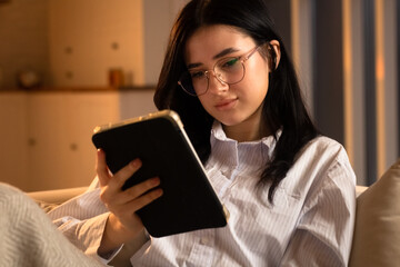 Cute woman in glasses uses a tablet while lying on the sofa. The long-haired brunette spends her evening watching TV shows on her digital tablet.