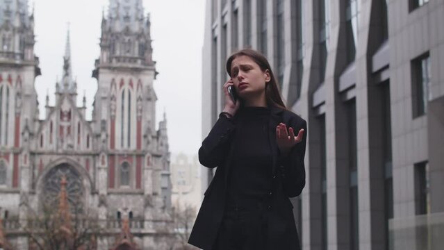 Upset young woman talking on the phone and gesturing on the street near the church