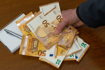 The hand of a man holding 50 euro bills on the background of the table and a notepad with a pen....