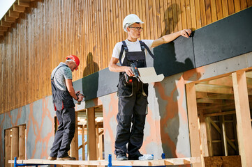 Carpenters constructing wooden framed house. Two men workers cladding facade of house with cement...