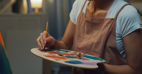 Close-up Youth Asia lady wear apron holding palette use brush painting on canvas create artwork cozy workshop in home at night. Contemporary Painter Abstract Modern Art, Creativity and people concept.
