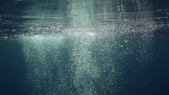 Motion of air bubbles rise up to surface in blue water in bright day sunlight, Slow motion. Sun beams penetrate water surface on bubbles rising underwater