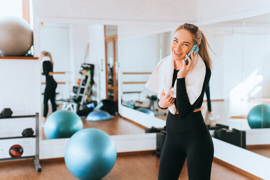 Excited sporty blonde young woman in sportswear and towel around neck talks by phone smiles stands at fitness class. Healthy lifestyle, sport. Caucasian female at break of training. Happy people.
