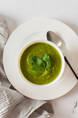 Healthy breakfasts, green broccoli and spinach cream soup in a white bowl on a marble background with a gray napkin, top view