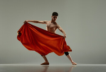 Dynamic image of handsome, artistic, young man, ballet dancer performing with red silk fabric over grey studio background. Art, classical dance, inspiration, creativity, beauty, choreography concept