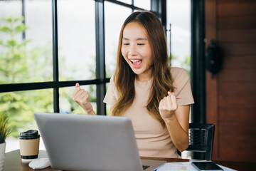 Excited happy Asian business young woman sitting with laptop computer at cafe desk raising hands up and celebrating success, female feeling winner rejoicing online win got new job at home
