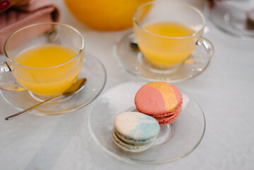 There are two macaroons in a transparent plate on a white table. Nearby are cups with orange juice...