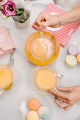 Cafe guests sit at a table and pour orange juice into glass cups. View from above. There are macaroons on the table in plates. There is a rose in a vase of water. Summer day