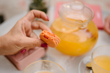 There are two macaroons in a transparent plate on a white table. Nearby are cups with orange juice and a teaspoon in a plate