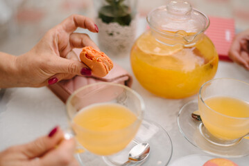 There are two macaroons in a transparent plate on a white table. Nearby are cups with orange juice and a teaspoon in a plate