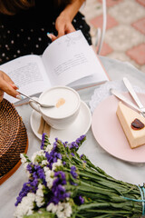 Summer cafe. On the table lies a woman's handbag woven from a wicker, a bouquet of wild flowers and an open book that the girl is reading. Dessert with strawberries on a plate. Spoon stirs coffee