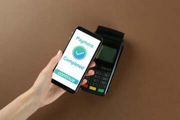 Woman using terminal for contactless payment with smartphone on brown background, top view. Transaction completed screen on device