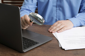 Man holding magnifier near laptop at wooden table, closeup. Online searching concept
