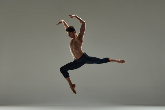 Young handsome man with muscular shirtless body, ballet dancer making performance over grey studio background. Concept of art, classical dance, inspiration, creativity, fashion, beauty, choreography