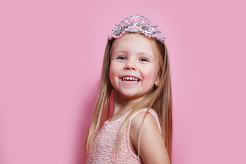 Close-up portrait of cute sweet little princess with toothy smile wearing light pink dress and diadem on pink background