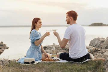 Couple having good time on date by lake sitting on rocky beach, laughing and clinking wine glasses....