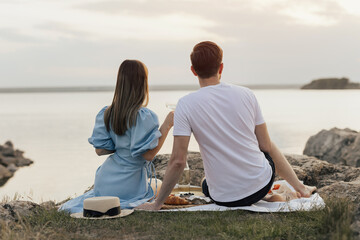 Happy young couple relaxing at the picnic. Guy and girl on a picnic drink white wine. Loving couple sitting on beach with picturesque landscape. 