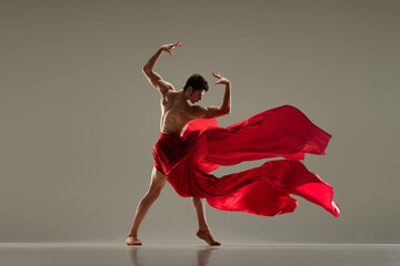 Fototapeta premium Handsome, attractive young man, professional ballet dancer dancing with red silk fabric against grey studio background. Concept of art, classical dance, inspiration, creativity, beauty, choreography