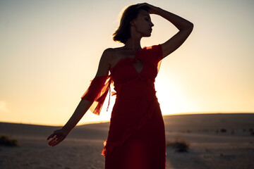 Portrait of a romantic woman in a red dress posing in the sand desert at sunset light.